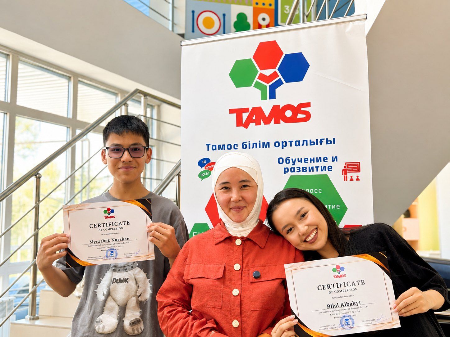 Подросток и две девушки у баннера Tamos, двое из них держат сертификаты об окончании курса русского языка